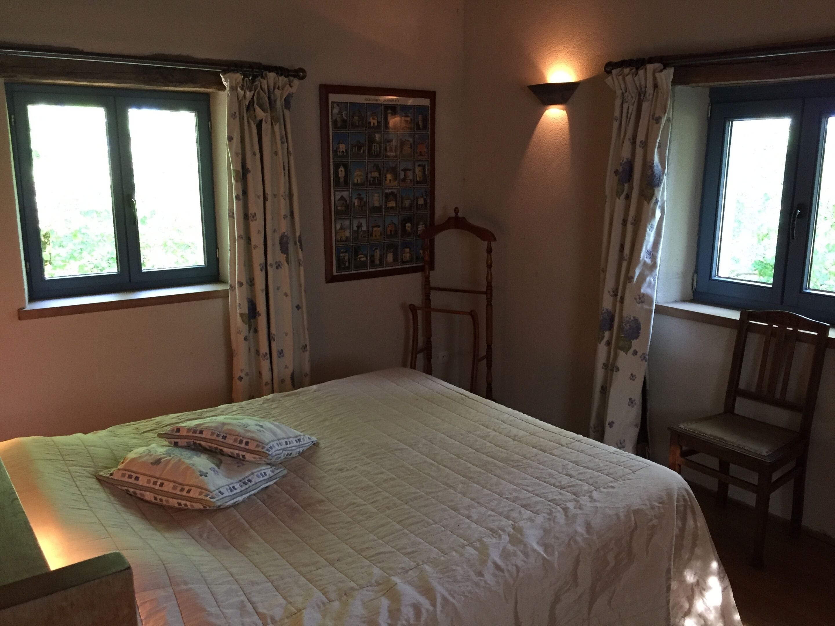 Un dormitorio con una cama, dos sillas, una silla de madera, un cuadro colgado en la pared y dos ventanas con cortinas.