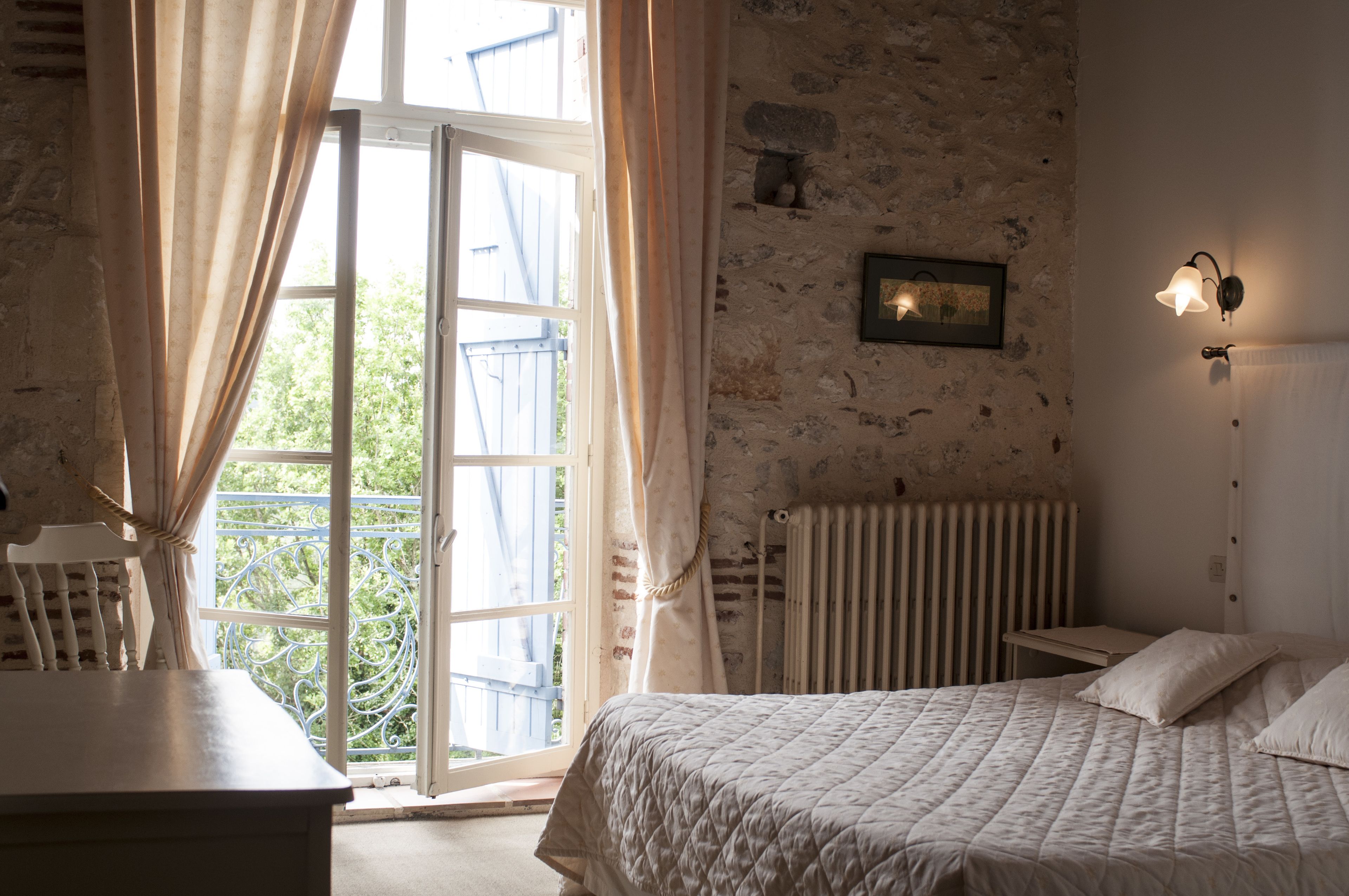 Une chambre à coucher comprenant un lit, un radiateur, une fenêtre avec des rideaux, une petite table et un luminaire fixé au mur.
