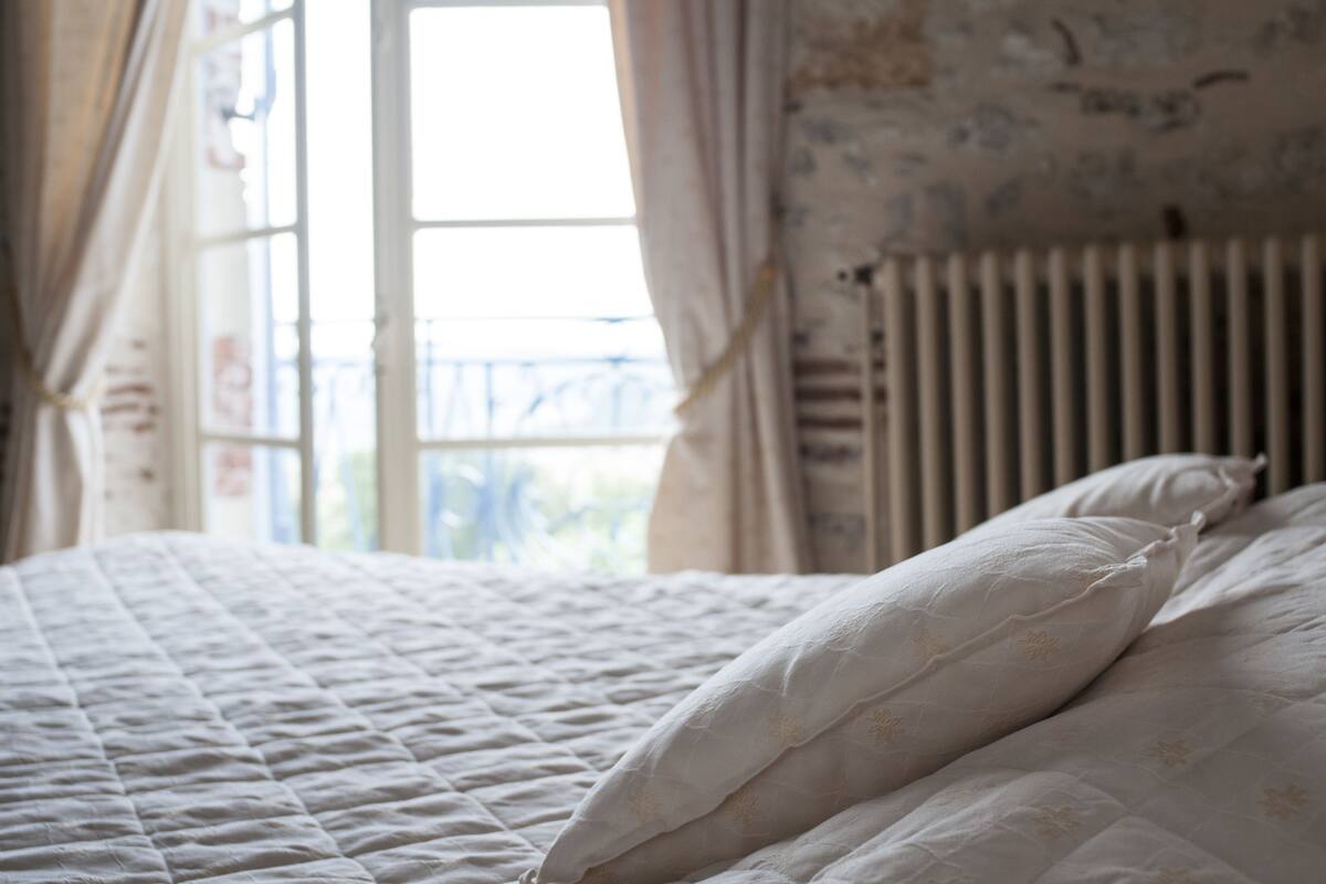 Ein Bett mit gesteppter weißer Bettdecke und Kissen, ein Fenster mit Vorhängen und ein Heizkörper.