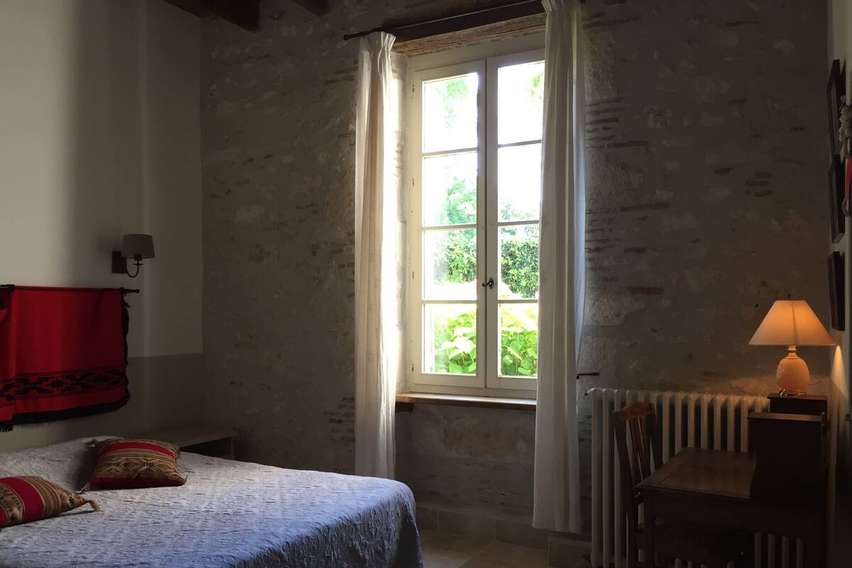 Ein Schlafzimmer mit einem Bett, einem Fenster mit Vorhängen, einem Heizkörper und einer Lampe auf einem Nachttisch.