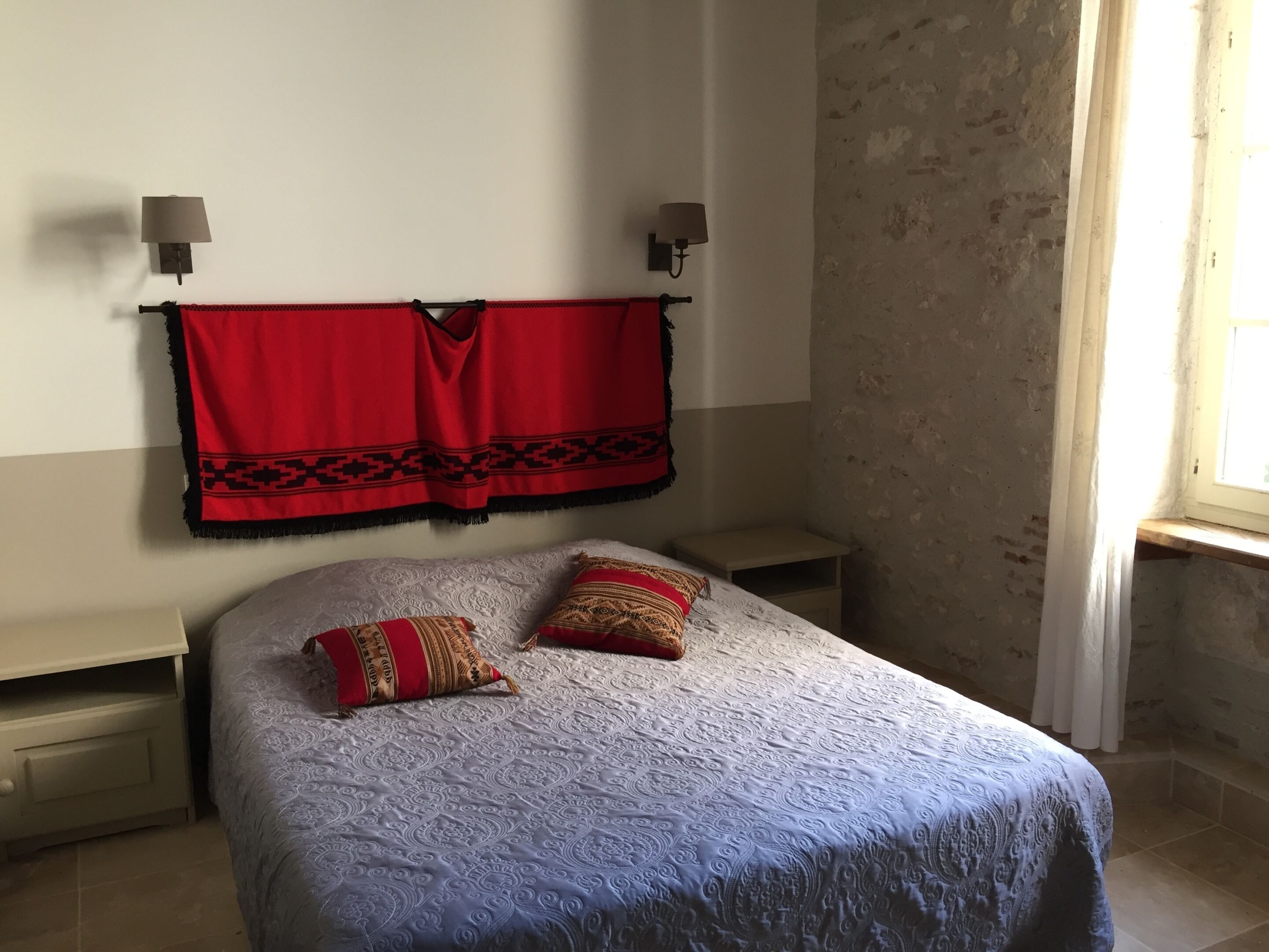 Quarto com cama, duas almofadas, manta preta e vermelha estampada, mesa de cabeceira e luminária na parede.
