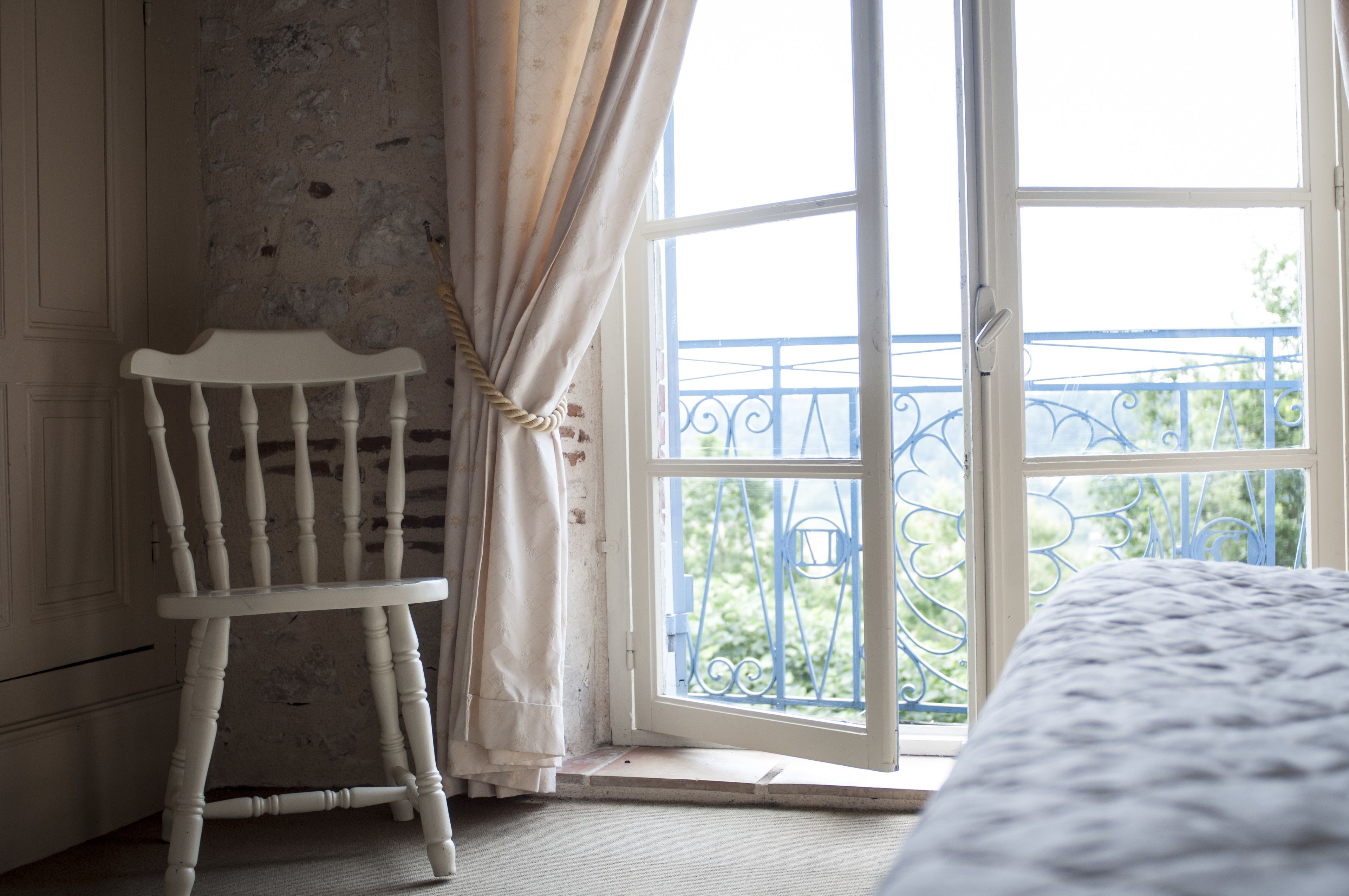 Une pièce avec une chaise blanche, une grande fenêtre donnant sur un balcon bleu et un lit recouvert d’une couverture à motifs.