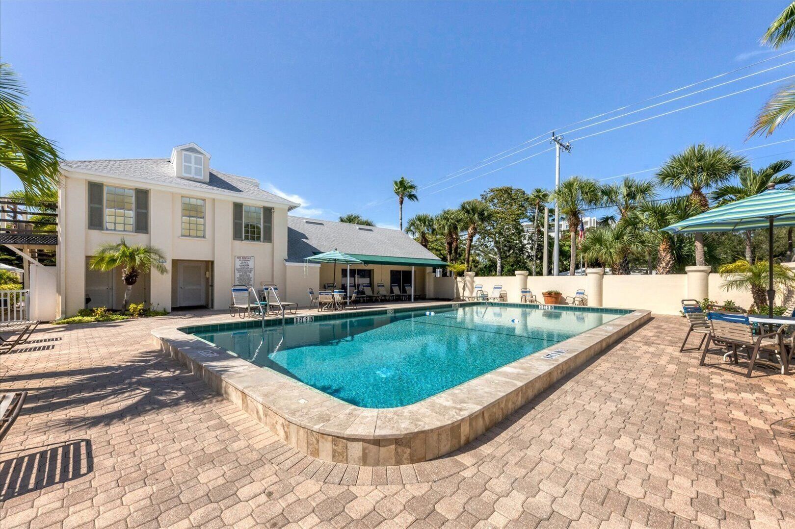 A swimming pool with lounge chairs and umbrellas, surrounded by a brick patio and palm trees.