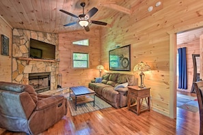 A cozy cabin interior with a stone fireplace, wooden walls, a reclining chair, a sofa, and a ceiling fan.