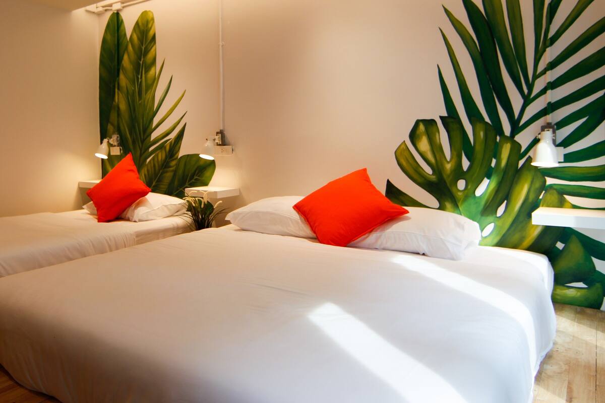 A room with two beds, each with white bedding and a red pillow. There are large green plants in the background, and the room has a wooden floor.