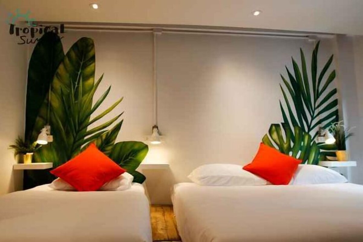 A hotel room with two beds, each with white bedding and a red pillow. There are large green plants on either side of the beds, and a small table with a lamp between them.