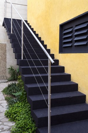 Una escalera con escalones negros y barandilla de metal que conduce a una pared amarilla con un ventanal con contraventanas negras.
