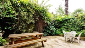 A wooden picnic table with benches and white metal chairs in a garden area with lush greenery and a stone wall. - Ladbroke Walk, by Veeve (London)