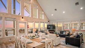 A spacious living area with a high ceiling, large windows, a dining table with chairs, and a seating area with sofas and a television. - Beachfront | Game Room, Hot Tub & Ocean Views (Bolivar Peninsula)