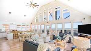 A spacious living area with a large sofa, a coffee table, and a television. There is a kitchen area with a dining table and chairs. The room has large windows offering a view of the ocean. - Beachfront | Game Room, Hot Tub & Ocean Views (Bolivar Peninsula)