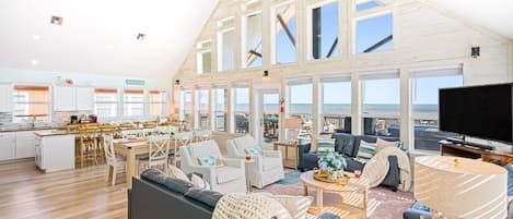 A spacious living area with a large sofa, a coffee table, and a television. There is a kitchen area with a dining table and chairs. The room has large windows offering a view of the ocean.