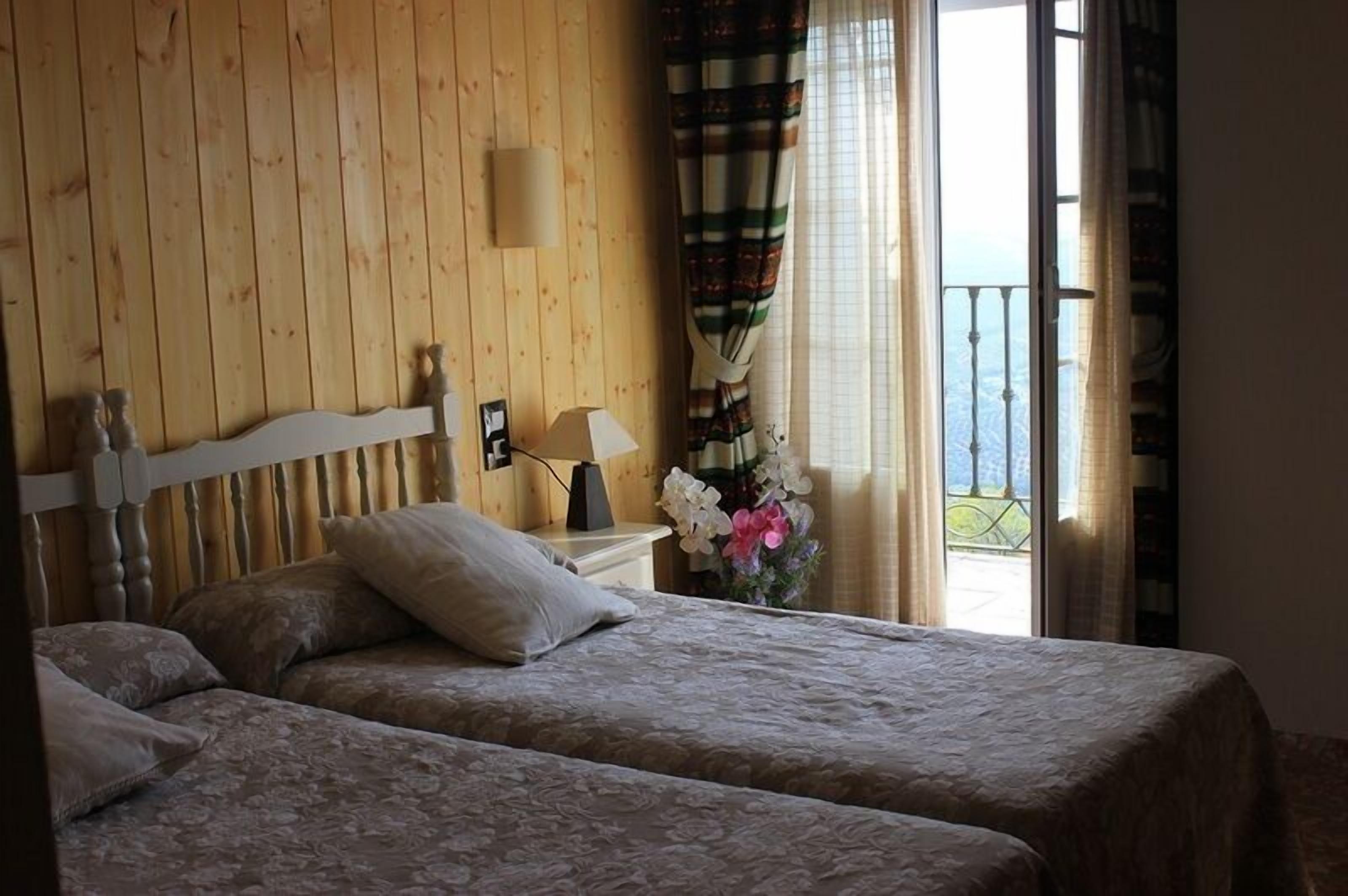 A room with two beds, a wooden headboard, a nightstand with a lamp, and a window with curtains.