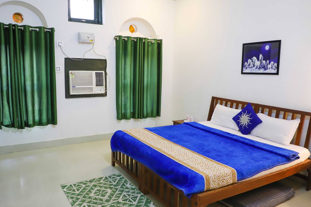 Quarto bem organizado com cama de madeira, cortinas verdes, ar condicionado fixado na parede, quadro emoldurado e tapete estampado.