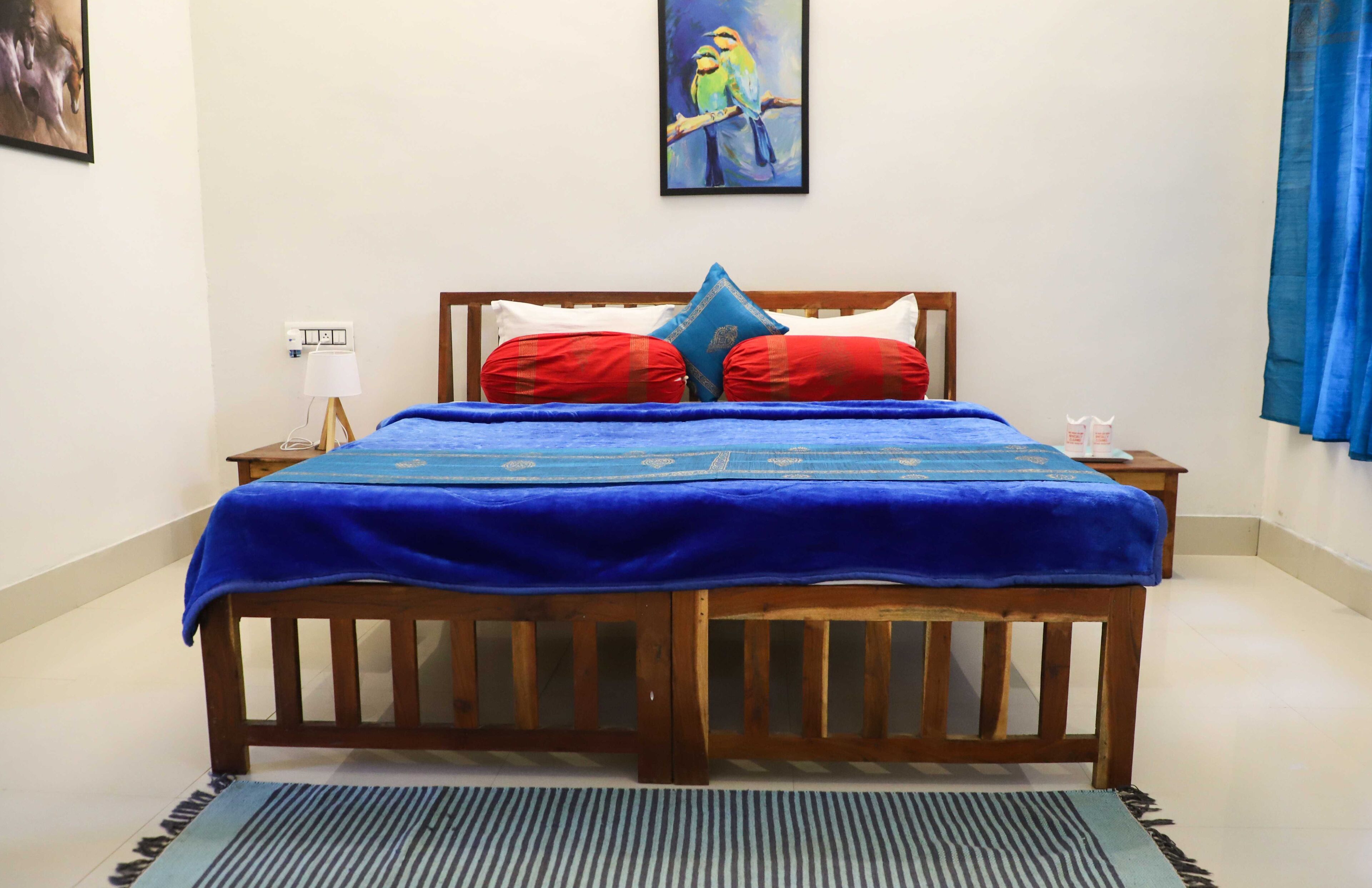 A neatly made bed with a blue bedspread, two red pillows, and a blue throw blanket. There is a framed painting of birds on the wall above the bed.