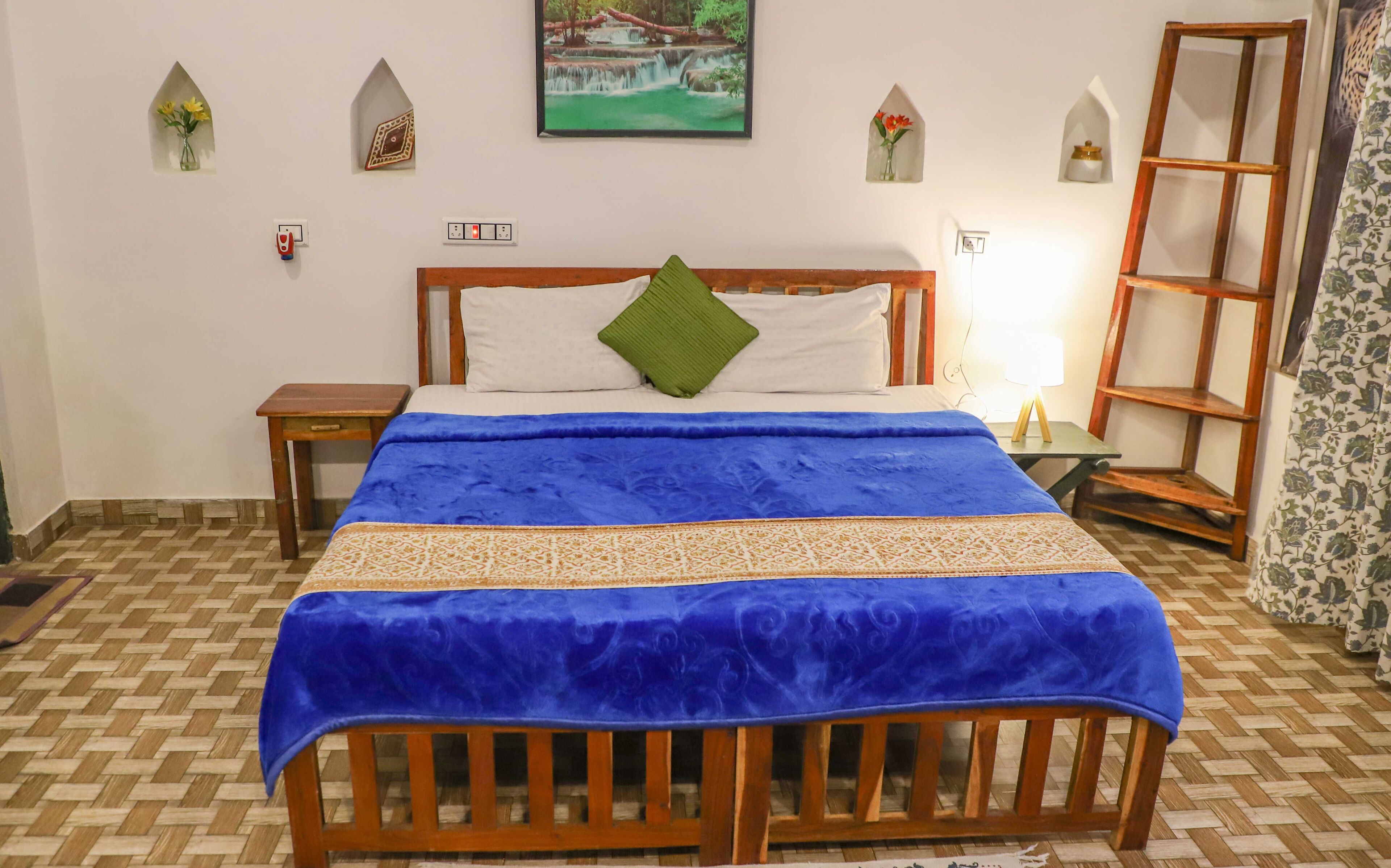 Una cama bien hecha con colcha azul, cabecera de madera y almohada verde. Hay dos mesitas de noche, una con lámpara, y una silla de madera con una bandeja.
