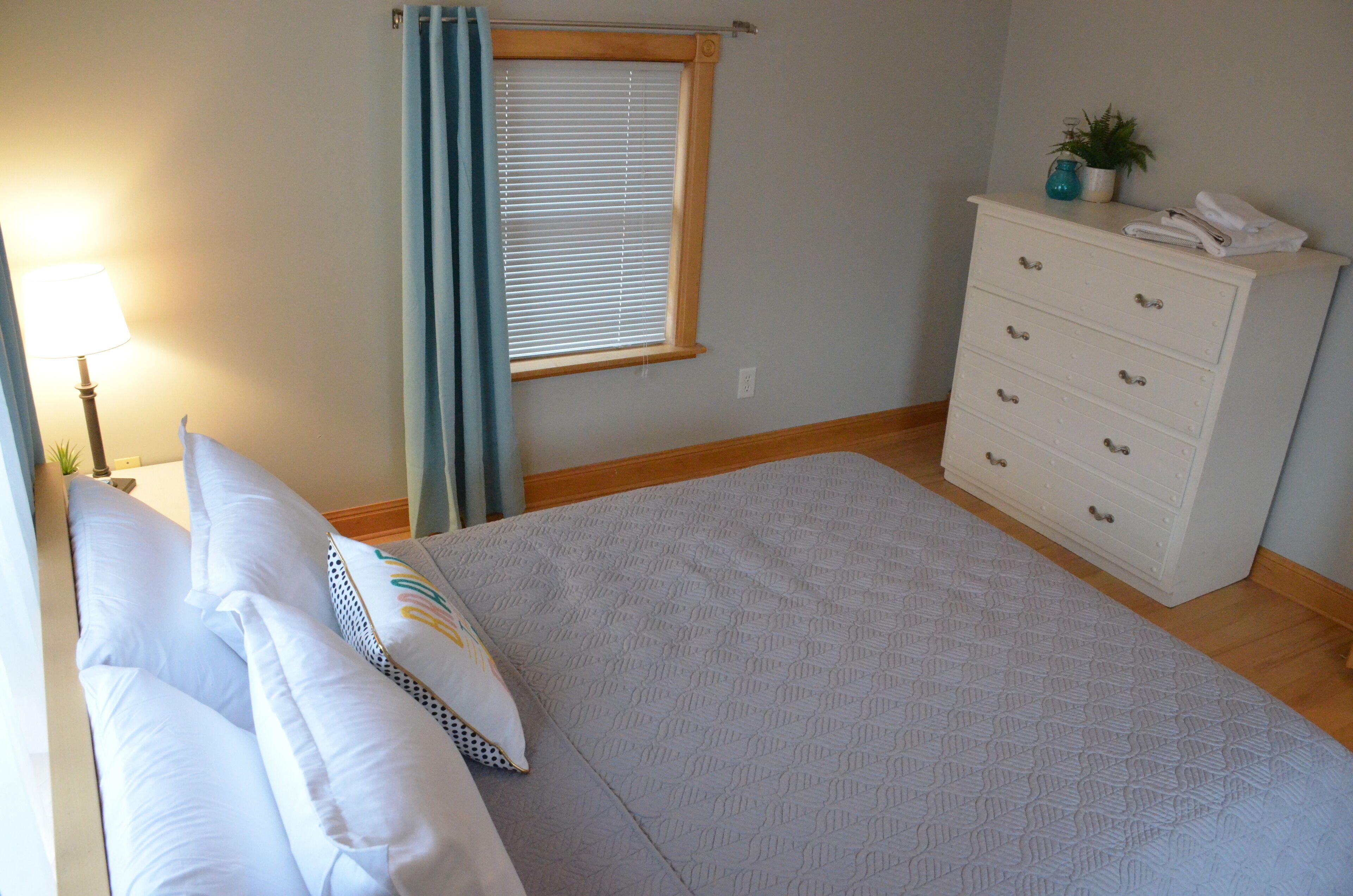 A bedroom with a bed, a dresser, a lamp, a window with curtains, and a small plant on the dresser.