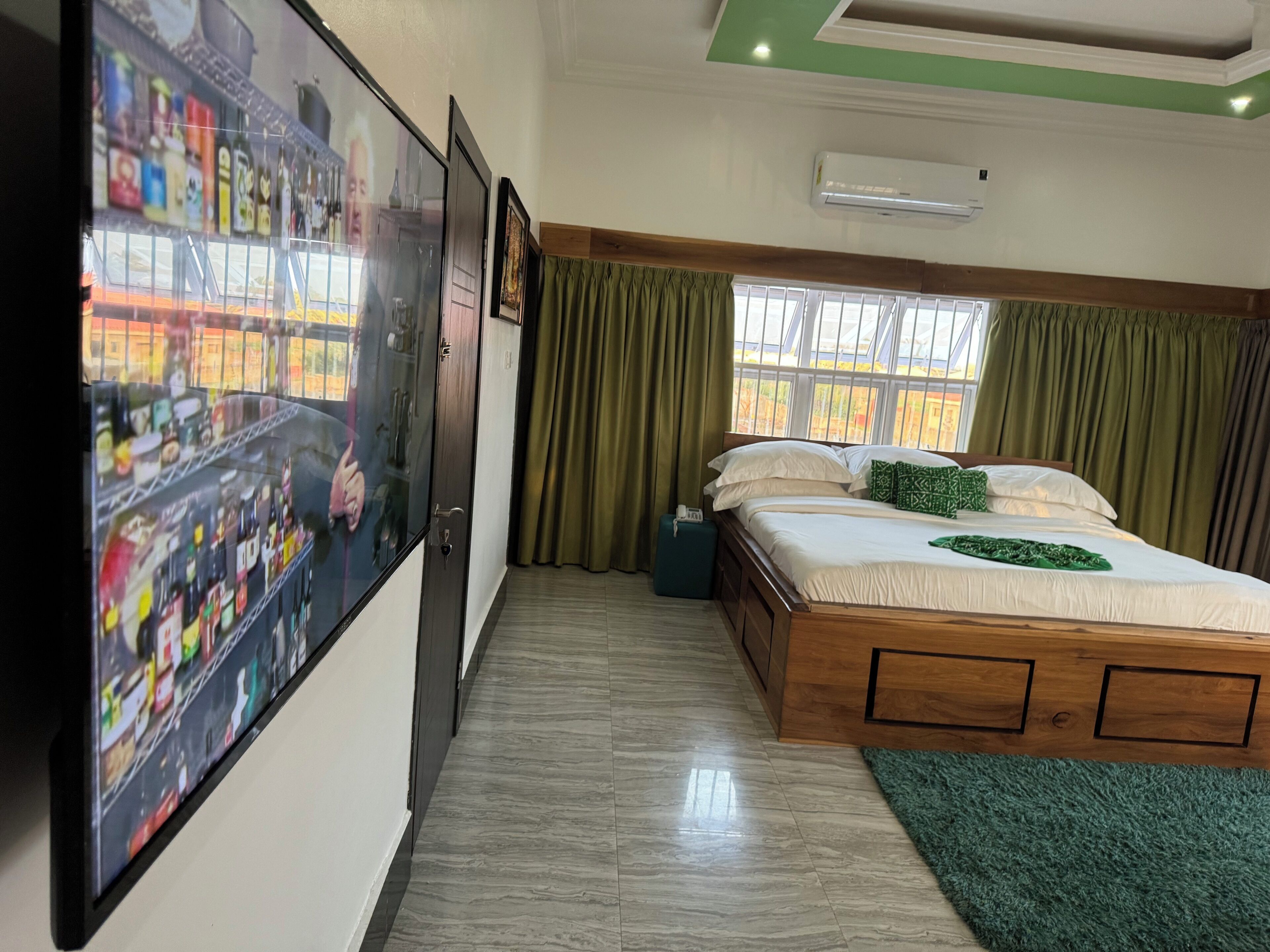 Una habitación de hotel con una cama, una alfombra verde, una ventana con cortinas, un aire acondicionado instalado en la pared y un televisor mostrando un paisaje urbano.