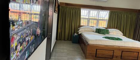 Una habitación de hotel con una cama, una alfombra verde, una ventana con cortinas, un aire acondicionado instalado en la pared y un televisor mostrando un paisaje urbano.