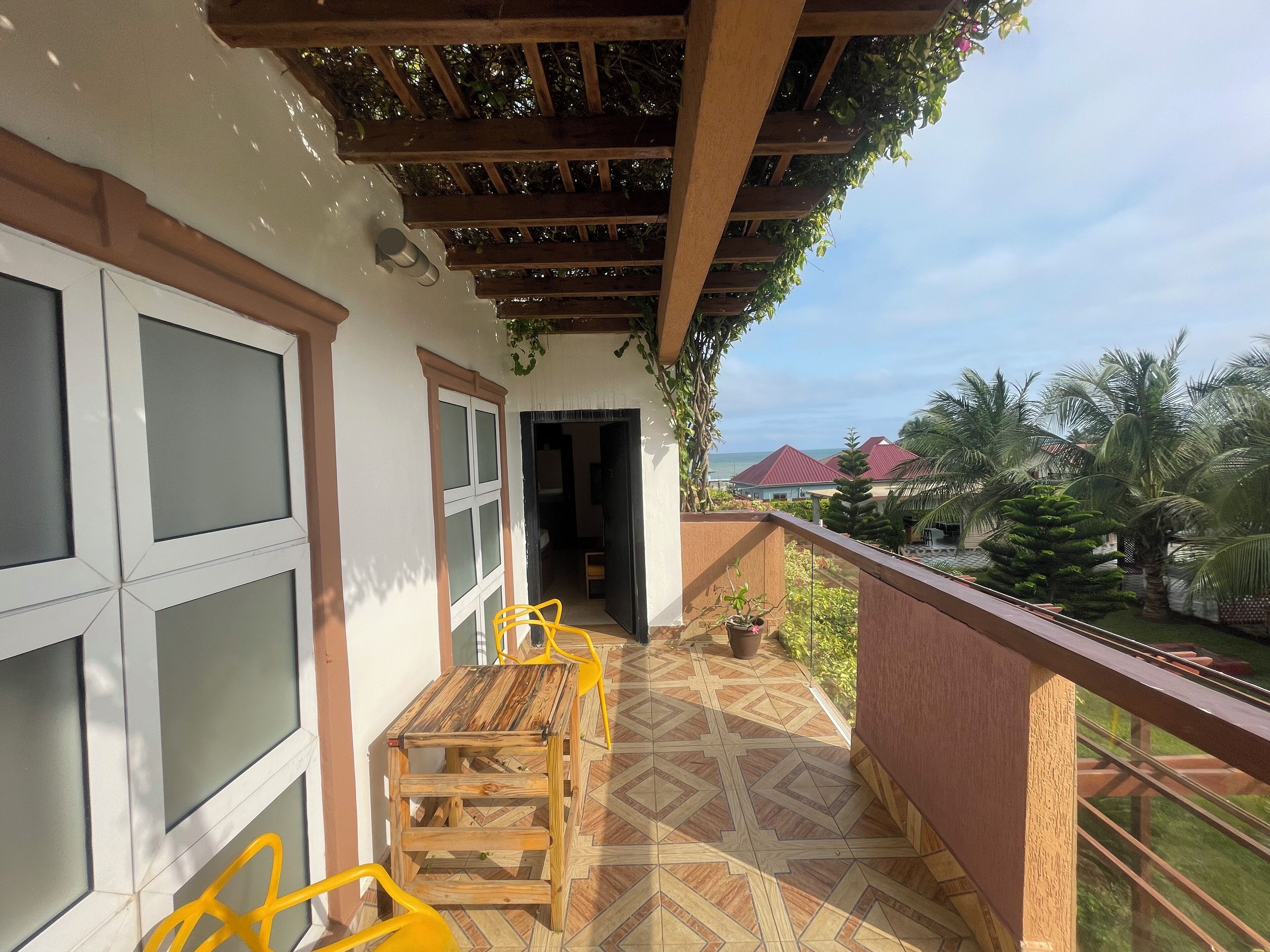 Un balcón con mesa y sillas de madera, vista al mar y un jardín con palmeras.