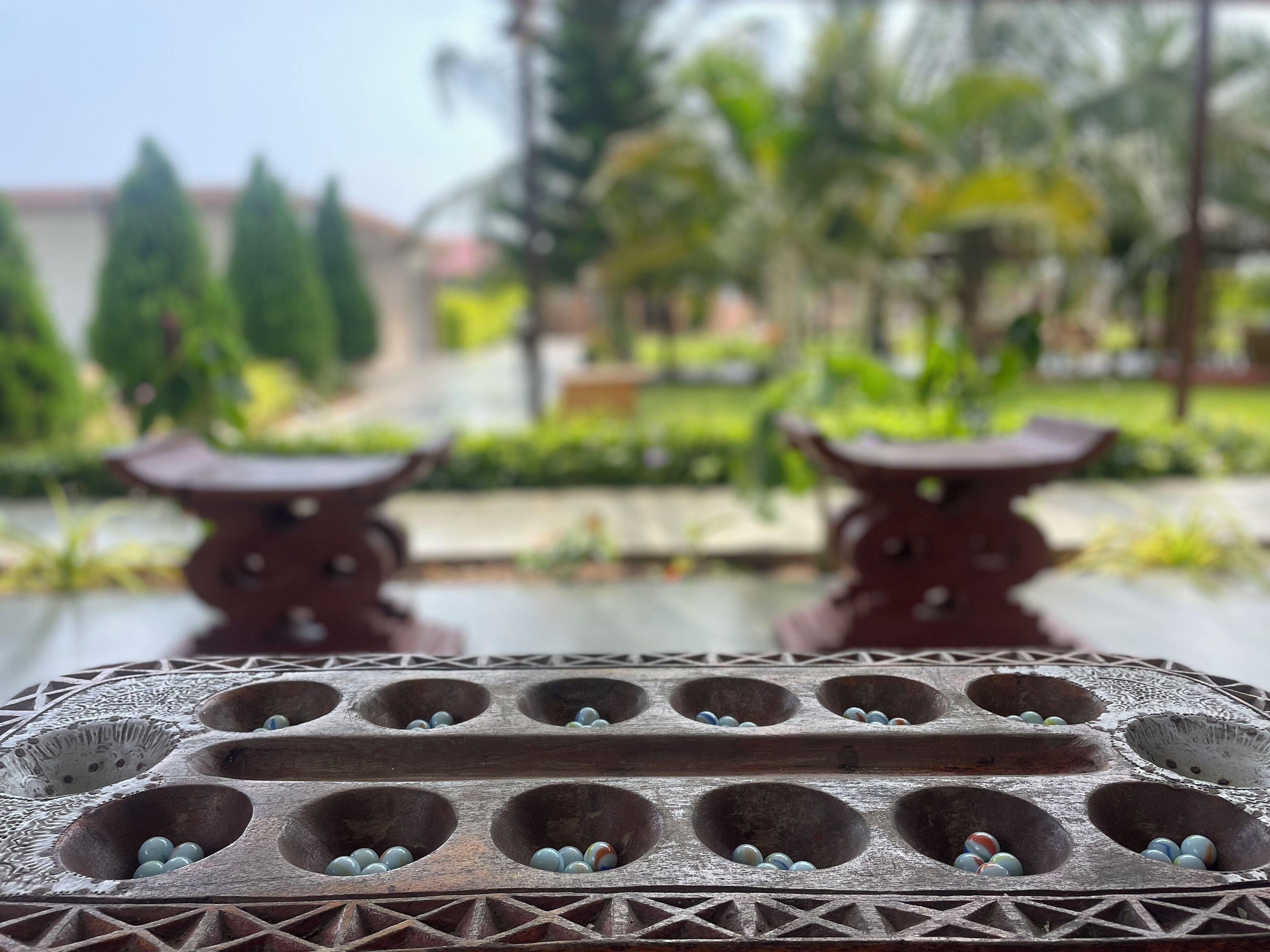 A close-up of a traditional wooden board game with multiple holes, set against a blurred outdoor background with greenery.