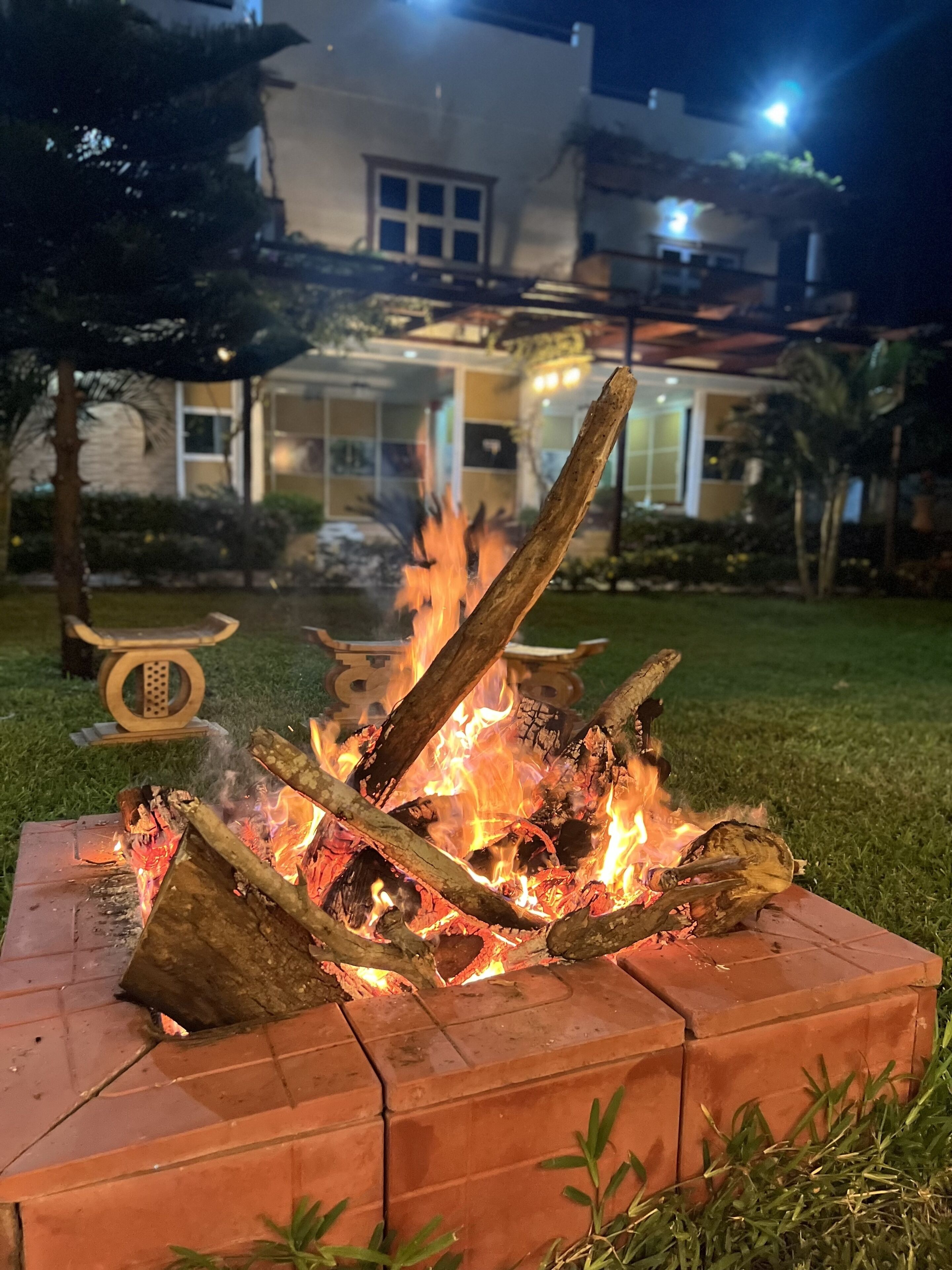 Uma lareira com lenha em brasa, em estrutura de tijolos, rodeada por grama e um jardim bem cuidado.