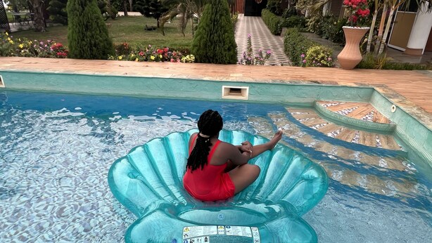 Una persona sentada en un flotador azul en una piscina con una terraza de azulejos y un jardín al fondo.