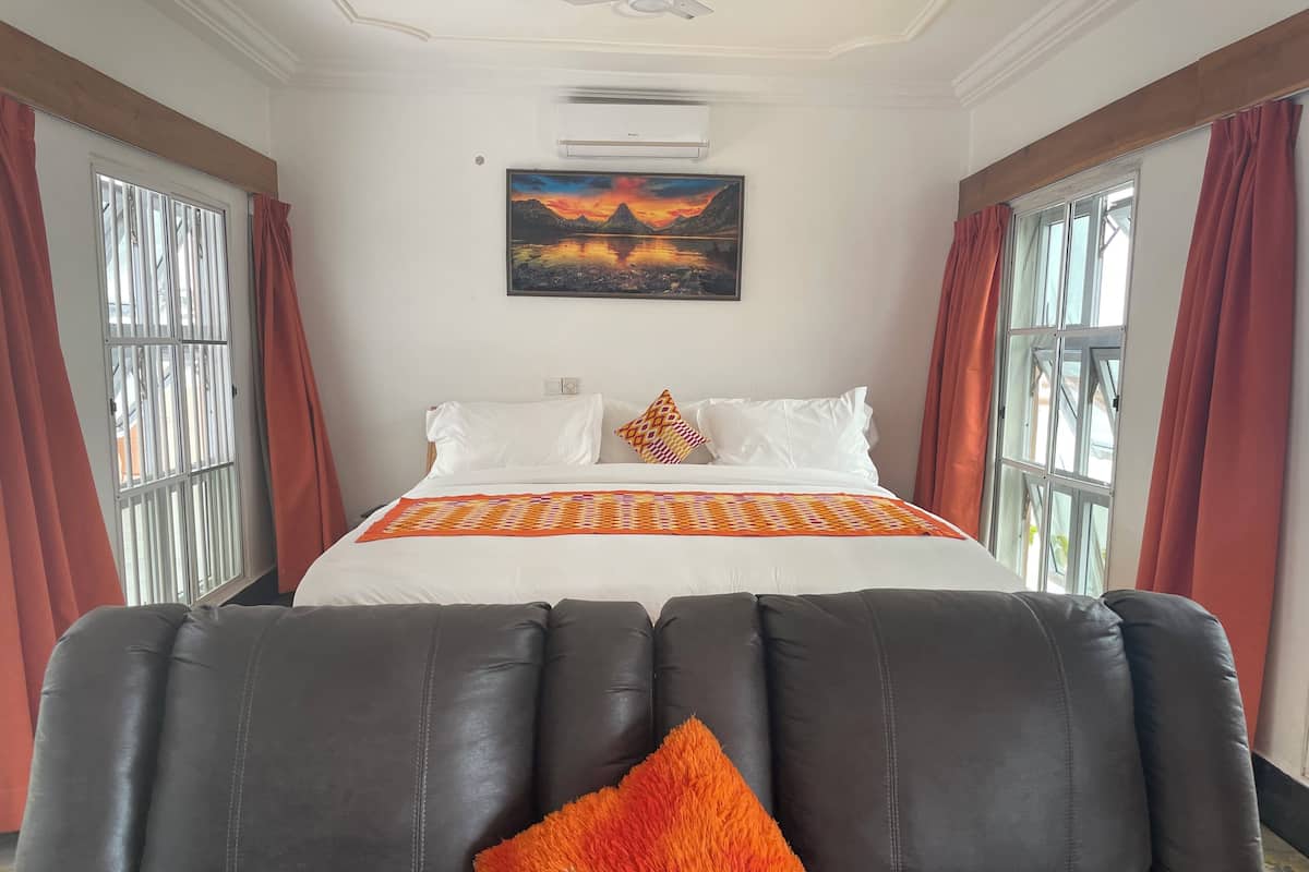 Quarto de hotel com cama, uma janela grande com cortinas, um ventilador de teto e um quadro na parede.