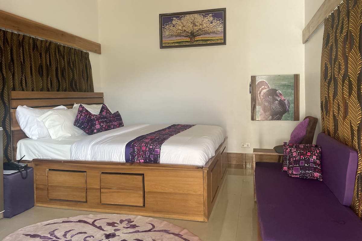 Um quarto com uma cama de madeira, um sofá roxo, um tapete estampado e duas molduras com quadros na parede.