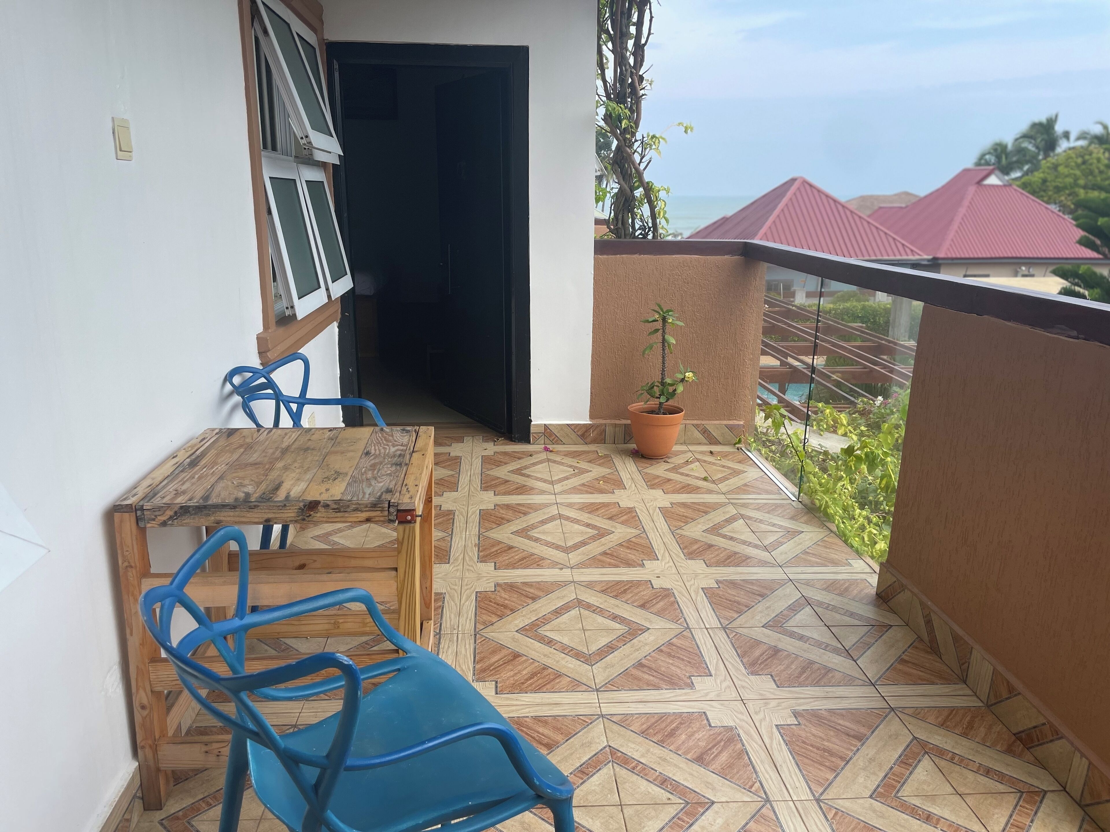A balcony with a wooden table and blue chairs, a potted plant, and a view of the sea and red-roofed buildings.