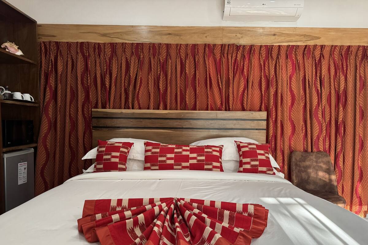 Uma cama bem arrumada com um cobertor estampado, cabeceira de madeira e cortinas vermelhas.