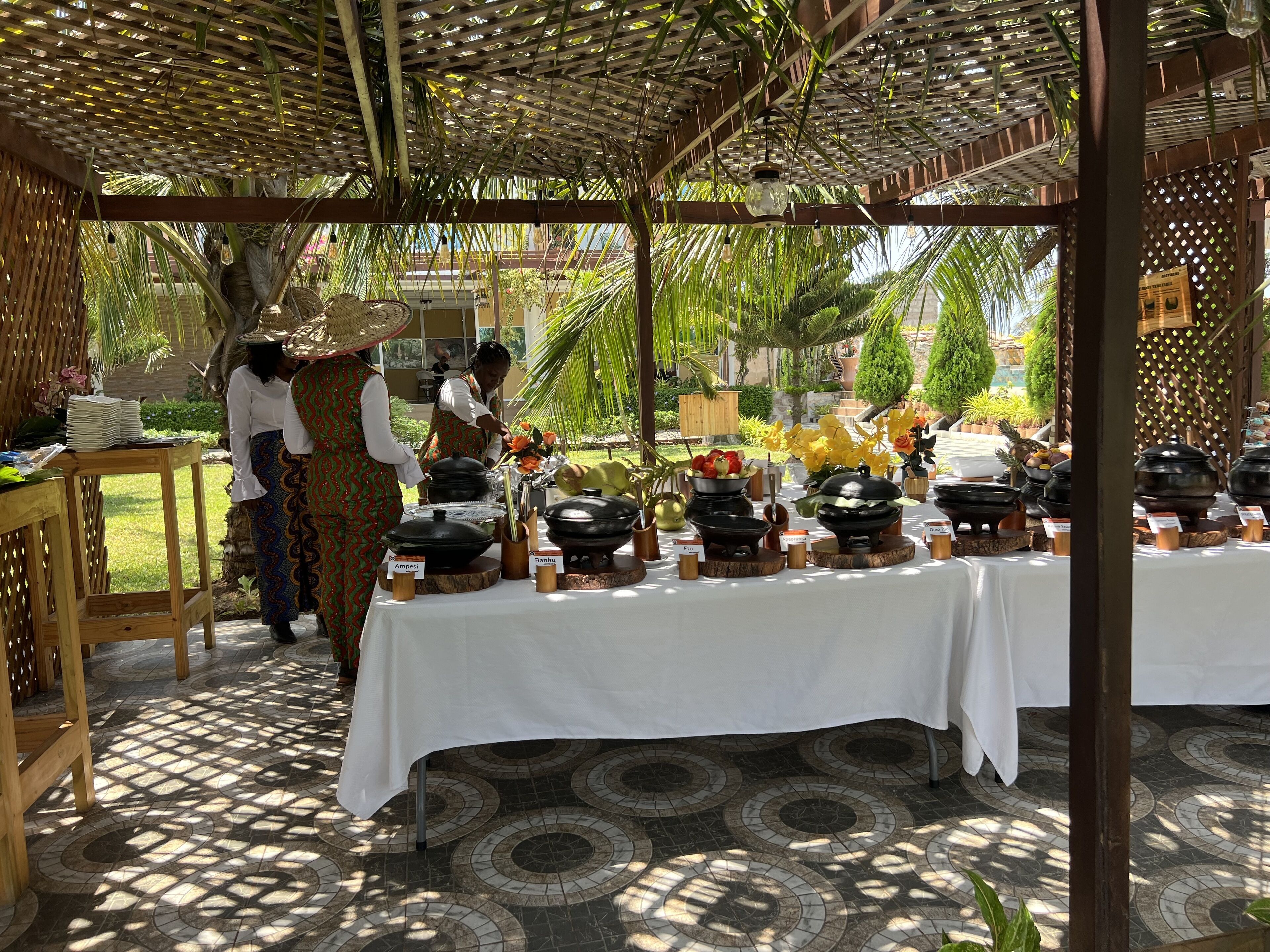 A covered outdoor dining area with a table set for a meal, surrounded by tropical plants and a pergola structure.