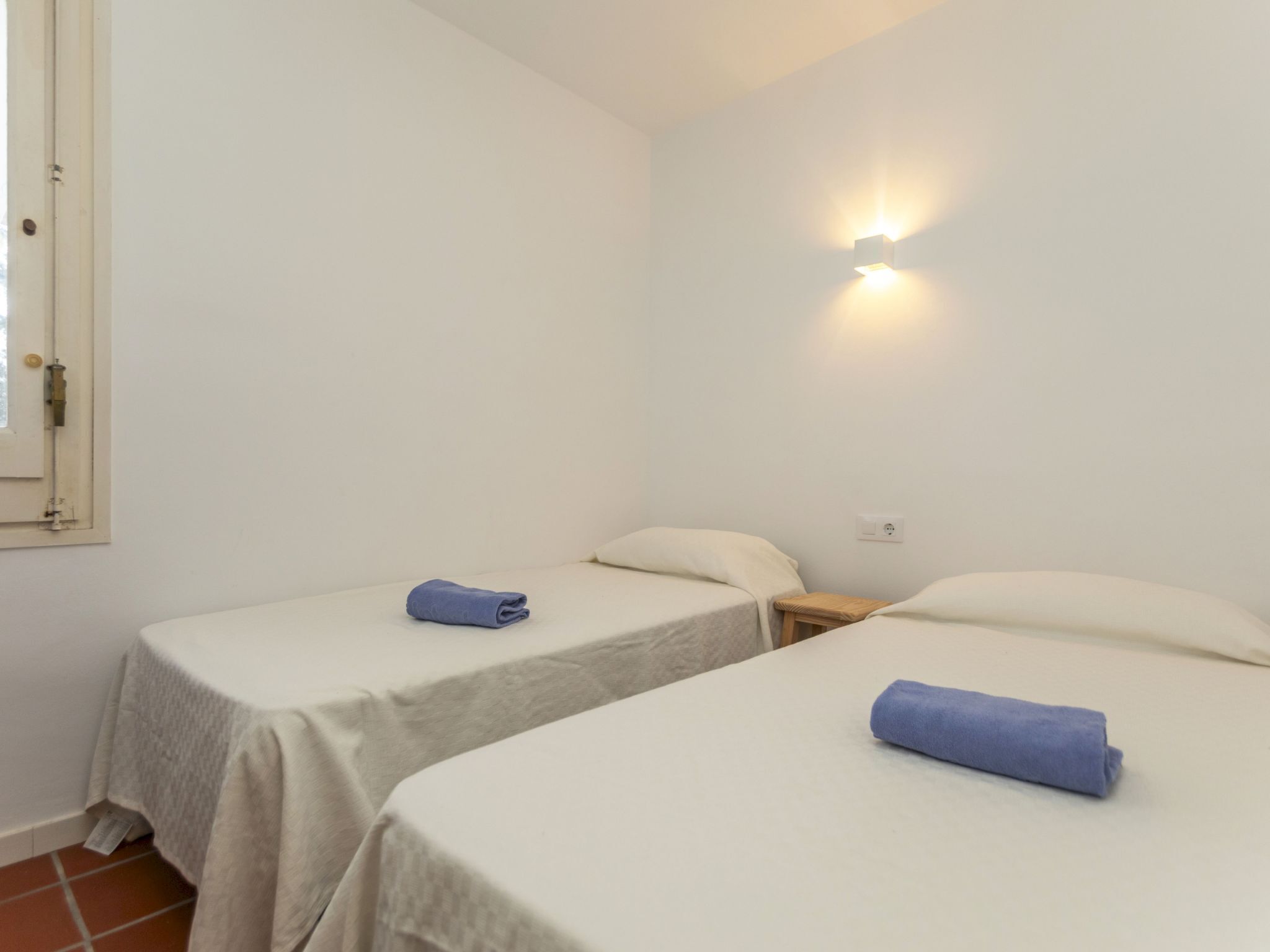 Una habitación con dos camas individuales, cada una con una colcha blanca y una toalla azul. Hay una mesita de noche de madera entre las camas, y una lámpara de pared sobre la cama de la derecha.