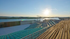 Ein leeres Freilichttheater mit terrassenförmig angelegten Sitzreihen, das auf ein Gewässer und ferne Hügel blickt.