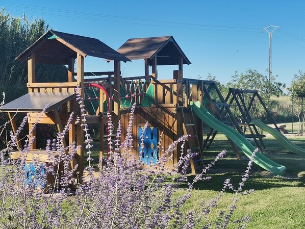 Une structure de jeu en bois, dotée de toboggans et d’éléments d’escalade, entourée de pelouse et de fleurs violettes.