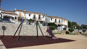 A playground with swings and a slide, surrounded by a stone wall and houses in the background. - Adosado Peñisol Inmomart (Peñiscola)
