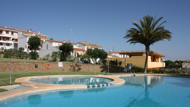 A swimming pool with a curved edge, surrounded by a stone wall and a palm tree. - Adosado Peñisol Inmomart (Peñiscola)