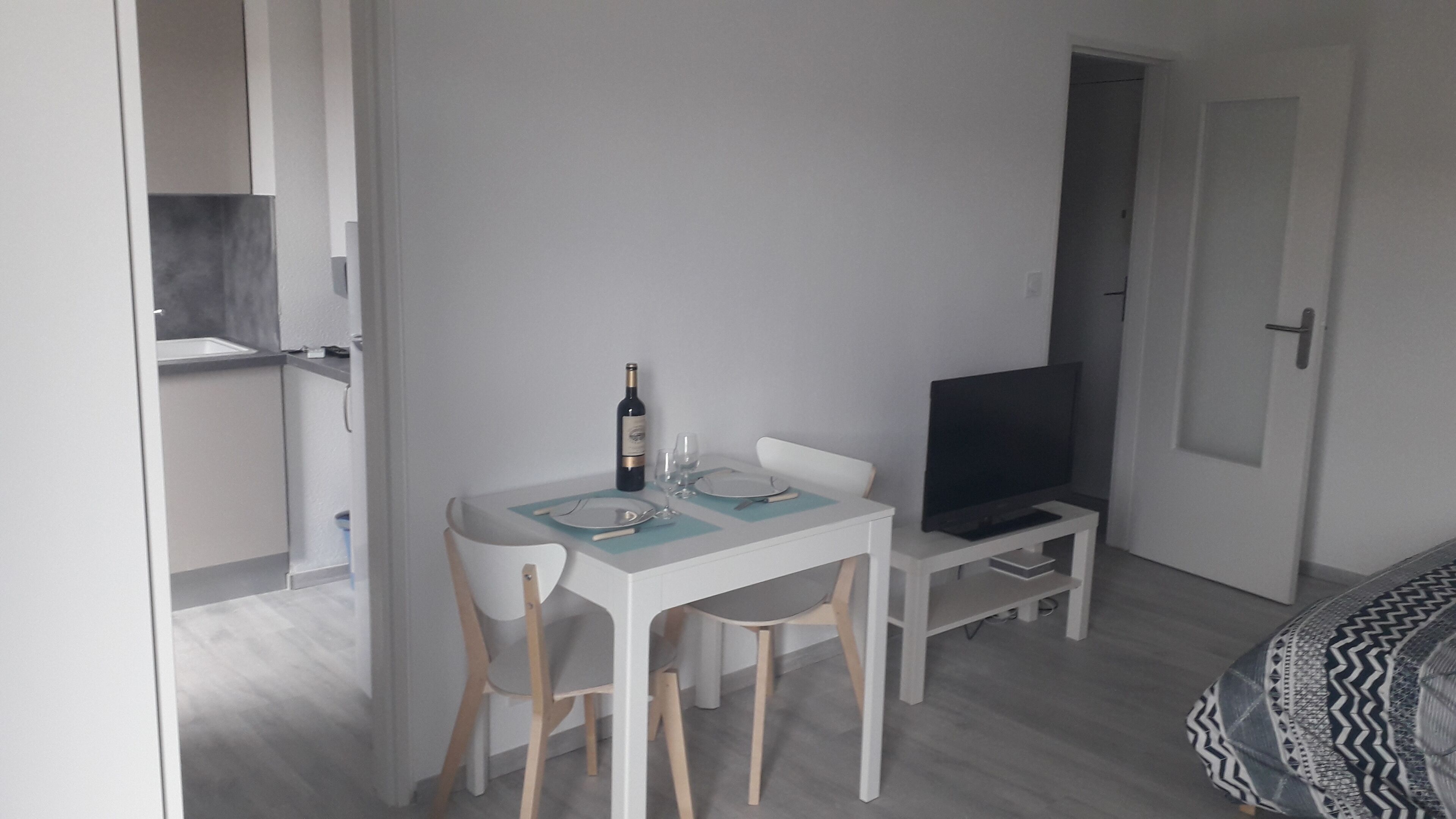 Una habitación pequeña y moderna con una mesa de comedor preparada para dos personas, un televisor sobre un mueble bajo y una puerta que da a otra habitación.