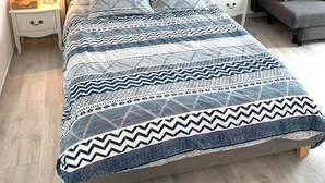 Sängyssä on kuviollinen päiväpeite, sen molemmin puolin on kaksi yöpöytää, joissa on lamput, huoneessa on harmaa sohva ja seinällä on taulu.