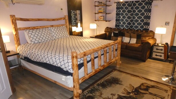 A wooden bed with a patterned bedspread, a wooden nightstand, a bedside lamp, a patterned rug, a brown leather sofa, and a wall-mounted shelf with books. - Yellowstone Studio # 3. Just One Mile From Yellowstone's Main Entrance (West Yellowstone)