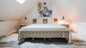 A bedroom with a sloped ceiling, a bed with patterned bedding, two bedside lamps, a painting on the wall, and a fluffy rug on the floor.