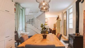 A modern kitchen with a wooden dining table, grey chairs, a hanging light fixture, and a staircase leading to another room.