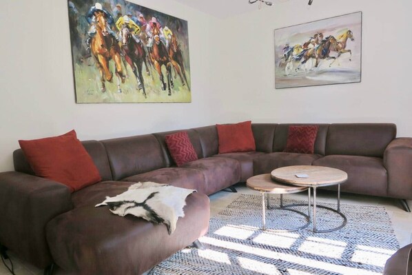 Un salon avec un canapé en cuir marron, des coussins rouges, une table basse ronde et deux tableaux accrochés au mur.