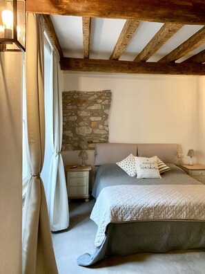 Une chambre avec un mur en pierre, un lit recouvert d’une courtepointe, une table de chevet avec une lampe et une fenêtre avec des rideaux.