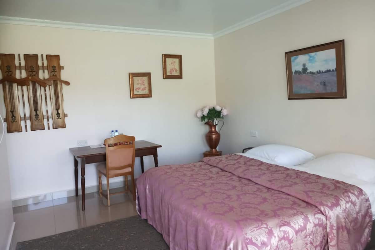 A hotel room with a bed, a desk, a chair, a vase with flowers, and two framed pictures on the wall.