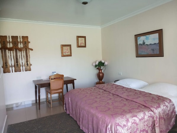 A hotel room with a bed, a desk, a chair, a vase with flowers, and two framed pictures on the wall.