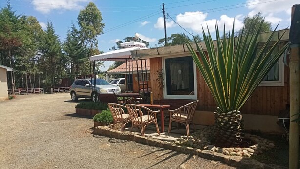 A small, rustic building with a covered outdoor seating area, a parked car, and a large agave plant. - Mlima View Gardens (Nanyuki)