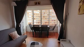 A compact living space with a sofa, a small table, a dining table with chairs, and a view of a building through large windows. - Studio Rosas Spain (ROSES)