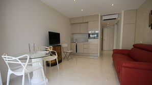 A modern living room with a glass dining table, white chairs, a red sofa, and a kitchen area with cabinets and appliances. - Modern Studio 1 minute from La Croisette (Cannes)