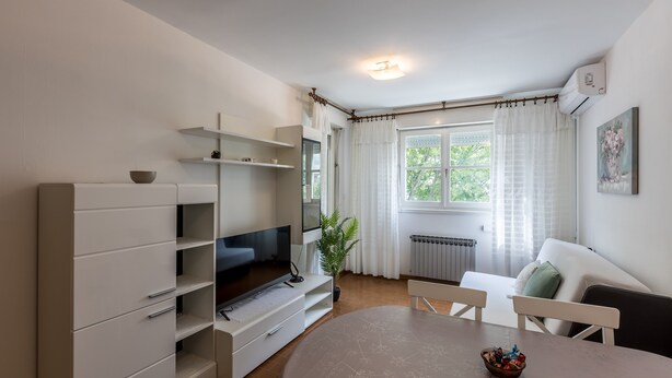 A modern living room with a flat-screen TV, white furniture, a dining table, and a window with curtains. - Apartment Gordana (ST) - One Bedroom Apartment (Split)