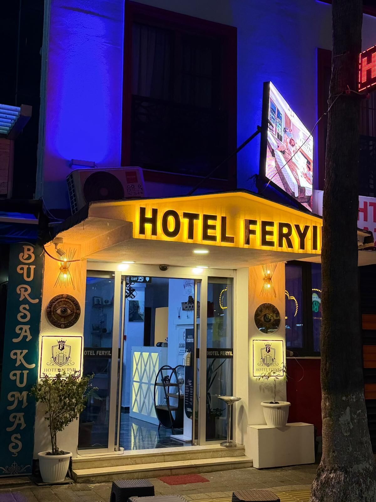 A hotel entrance with a sign reading 'HOTEL FERYI,' a potted plant, and a chair outside.